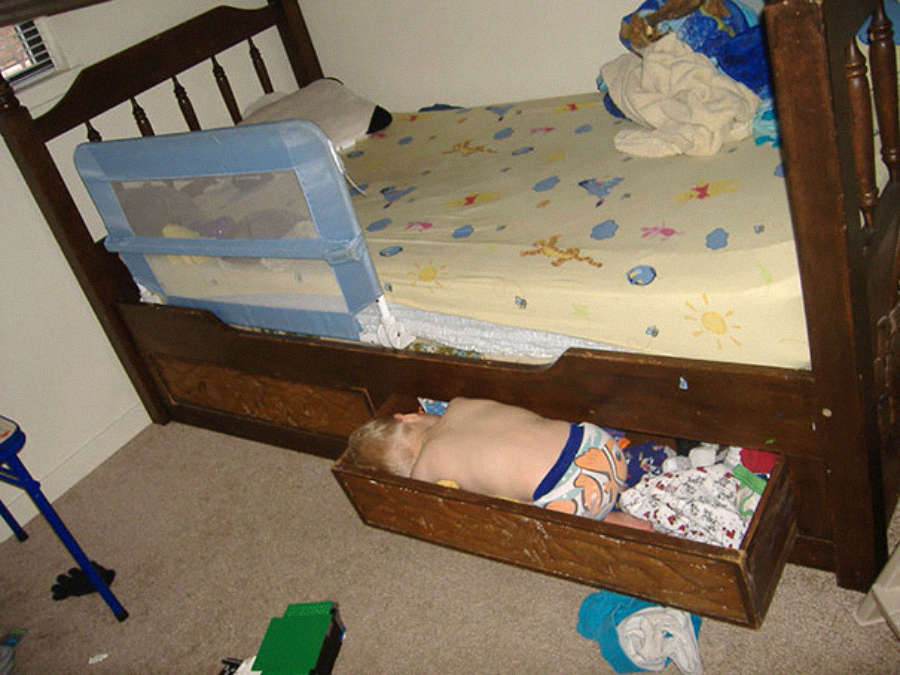 A small child wearing cartoon-printed pajamas is sleeping inside an open wooden dresser drawer underneath a bed, surrounded by scattered clothes and toys on the carpeted floor.