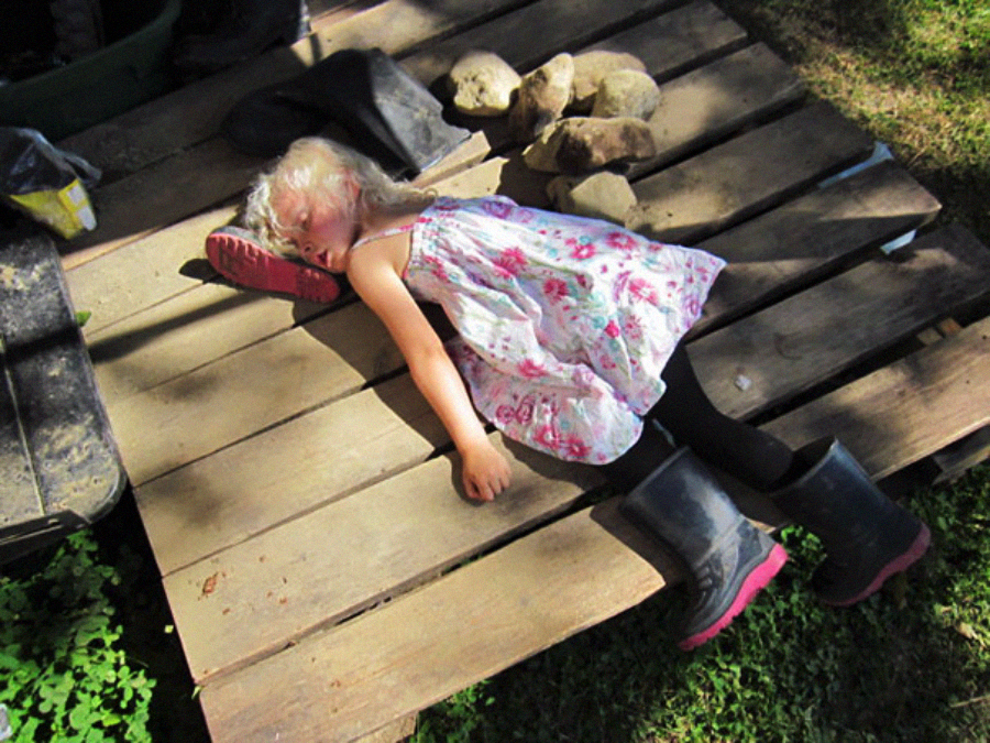A young girl in a floral dress and black boots is lying on her side on a wooden pallet outdoors, appearing to rest or nap in the sunlight, with rocks and shadows nearby.