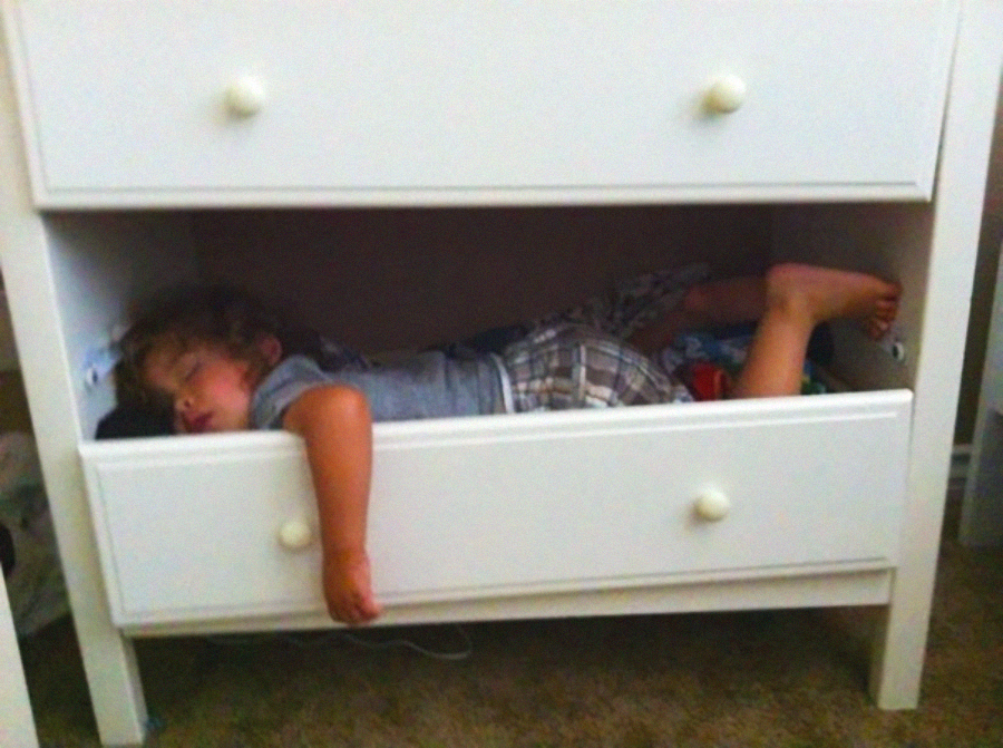 A young child is sleeping inside an open drawer of a white dresser, with their head resting on clothes and one arm and leg hanging out.