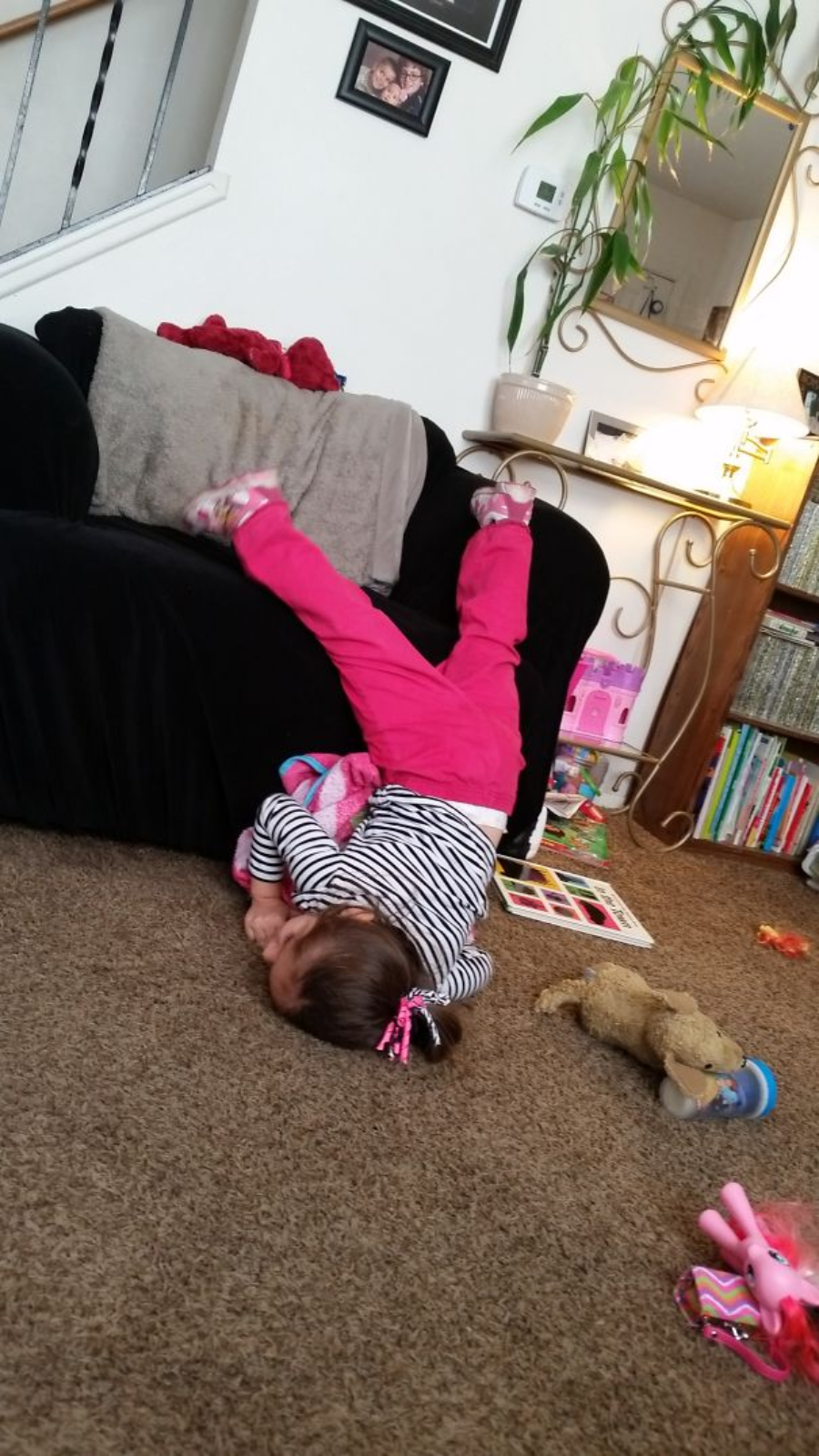A young child in pink pants and a striped shirt lies upside down on the floor with legs up on a black couch in a cozy living room, surrounded by toys and books.