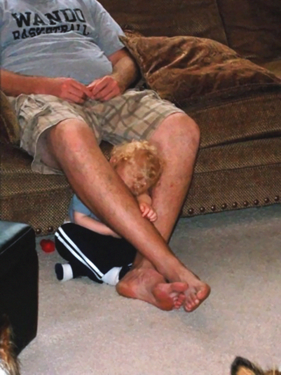 A small child with curly blonde hair sits on the floor, leaning their head against an adult’s bare legs. The adult, wearing shorts and a Wando Basketball shirt, sits on a brown couch.