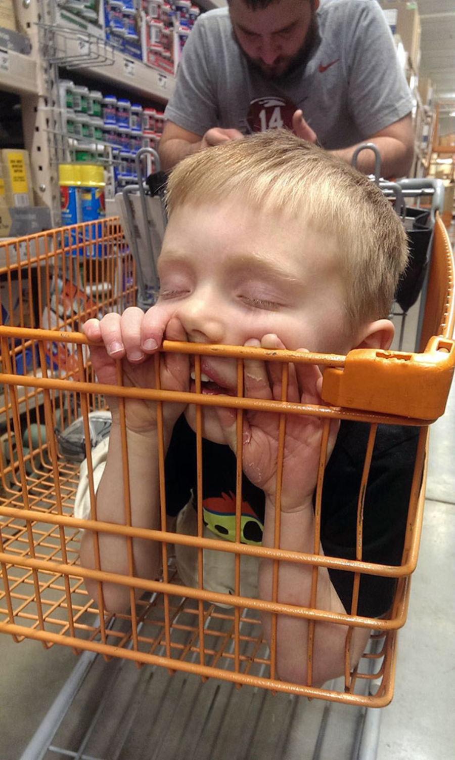 A young child is asleep with their face resting against the side of an orange shopping cart, eyes closed and mouth slightly open. An adult stands behind the cart in a store aisle.