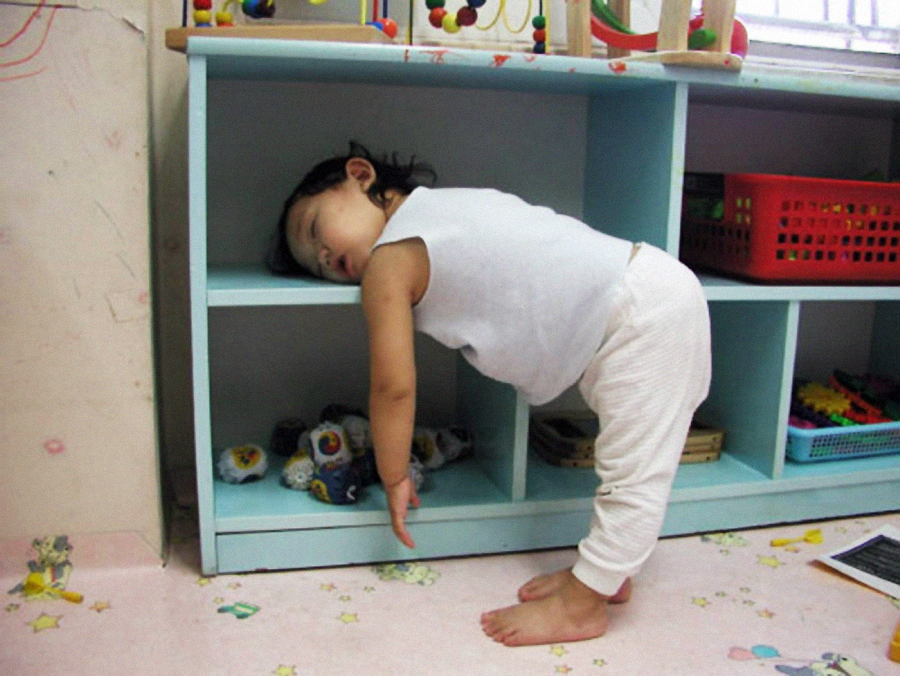 A toddler wearing white clothes is asleep while standing, leaning face-first onto a blue shelf with their arms hanging down and toys visible in the background.