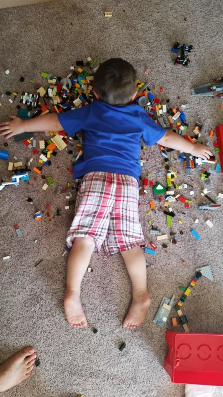 A young boy in a blue shirt and plaid shorts lies face down on a carpet, arms outstretched, surrounded by scattered colorful LEGO bricks and pieces.