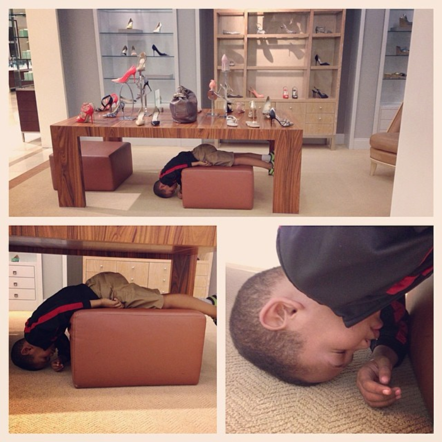 A young boy lies upside down and crouched under a wooden display table in a shoe store, appearing bored as shoes and handbags are displayed above him. Two close-ups show him resting his head on the floor, eyes closed.