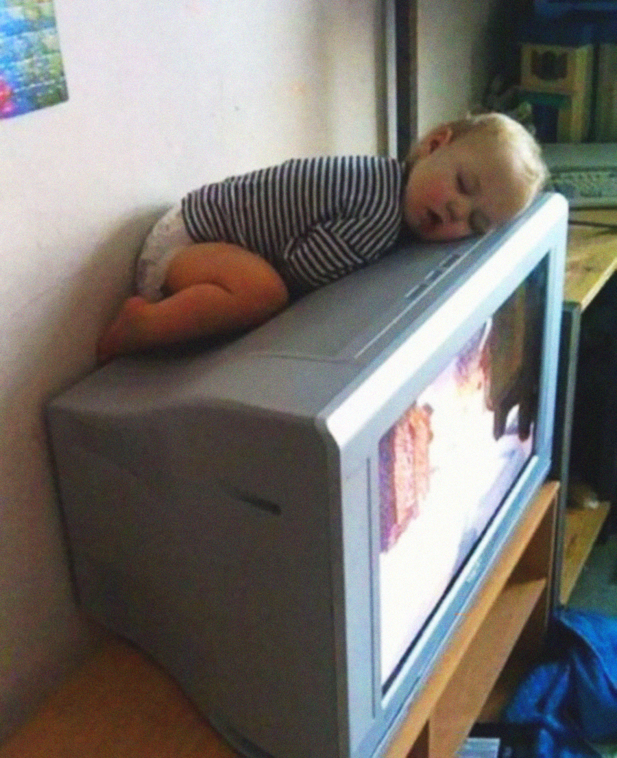 A toddler in a striped shirt and diaper sleeps on top of an old box-style television, using it as a pillow while the TV is turned on, showing an image on the screen.