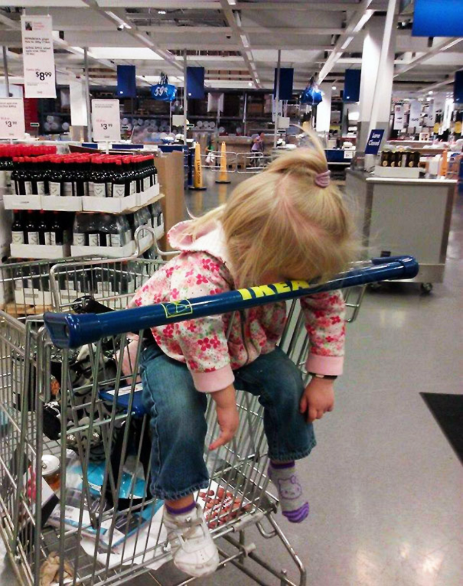 A young child is slumped over asleep inside a shopping cart in a store, with her head resting on the blue handlebar. She wears jeans, a floral jacket, and sneakers. Shelves and bottles are visible in the background.