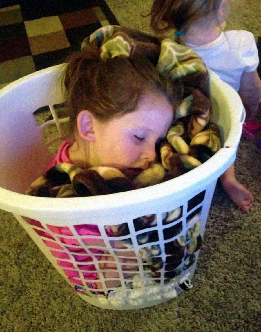 A young child sleeps curled up in a white laundry basket, wrapped in a patterned blanket. Another child sits nearby on a carpeted floor with her back to the camera.