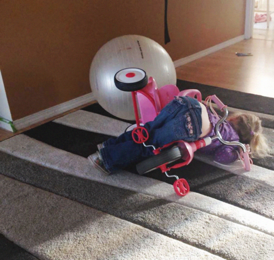 A child lies face down on a striped carpet after falling off a small pink tricycle, with a large exercise ball nearby. The tricycle is tipped over on its side.