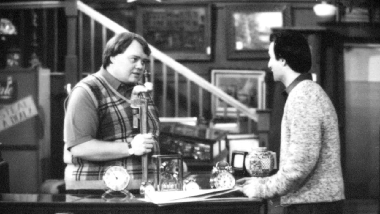 Two men talk at a counter in a shop filled with clocks and antiques. One holds a lamp, while the other listens. Framed pictures and a staircase are in the background. The image is in black and white.