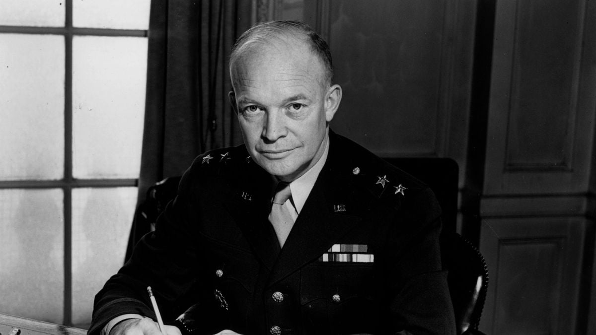 A man in a military uniform with medals sits at a desk, holding a pen, looking directly at the camera. There is a window and curtains in the background.
