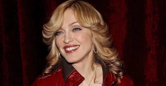 A smiling woman with wavy blonde hair, wearing a red leather jacket and hoop earrings, stands in front of a deep red curtain.