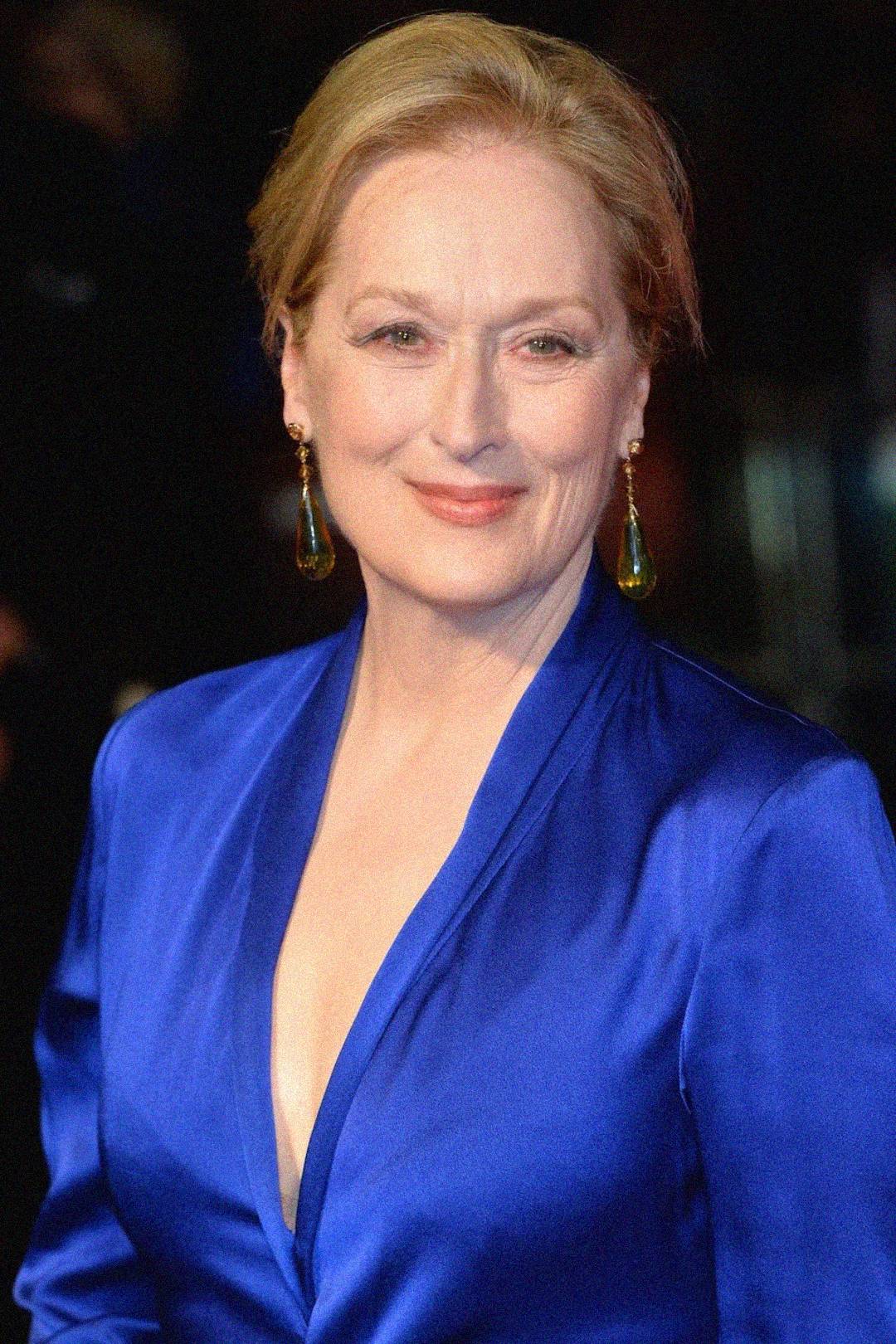 A woman with light skin and blonde hair is smiling and wearing a royal blue dress with a deep neckline and long sleeves. She also has drop earrings. The background is dark and blurred.