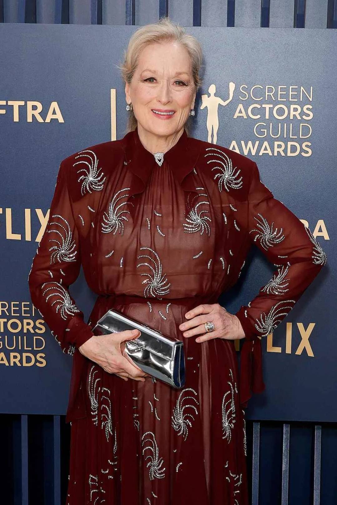 A woman in a maroon dress with silver swirl designs smiles at the 2023 Screen Actors Guild Awards, posing with one hand on her hip and holding a silver clutch, standing in front of a blue backdrop with event logos.