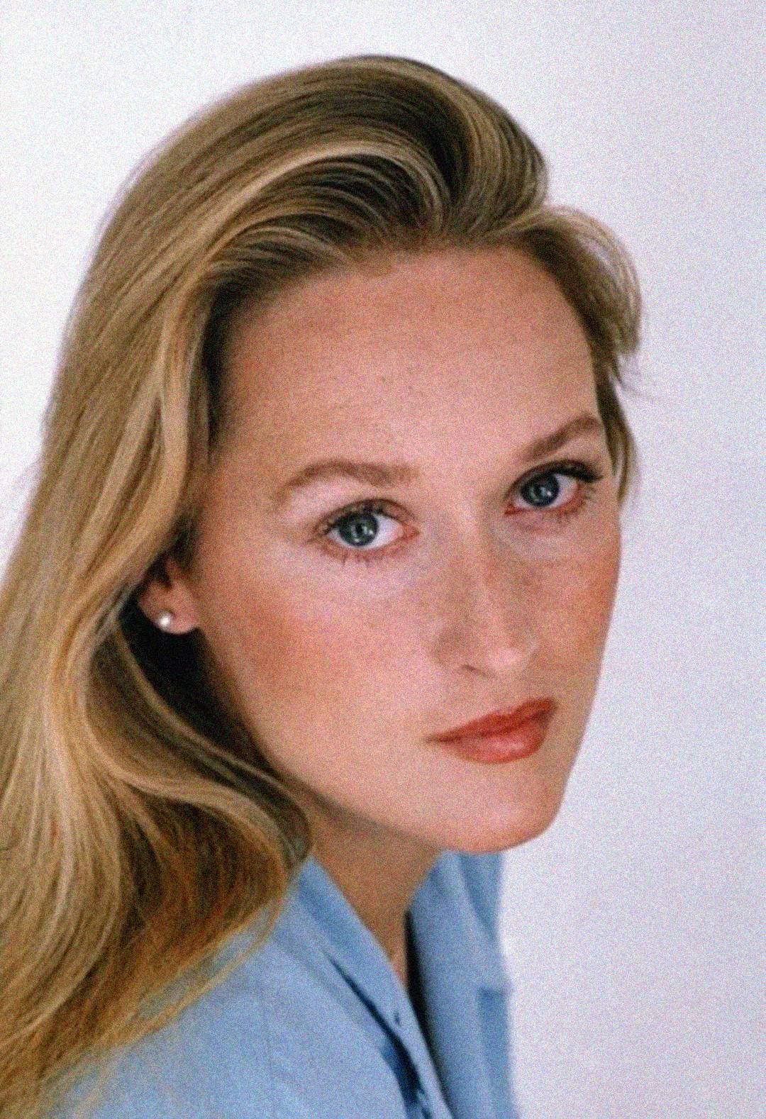 A woman with long blonde hair, light skin, and blue eyes gazes at the camera with a neutral expression. She is wearing a light blue top and small stud earrings, set against a plain white background.