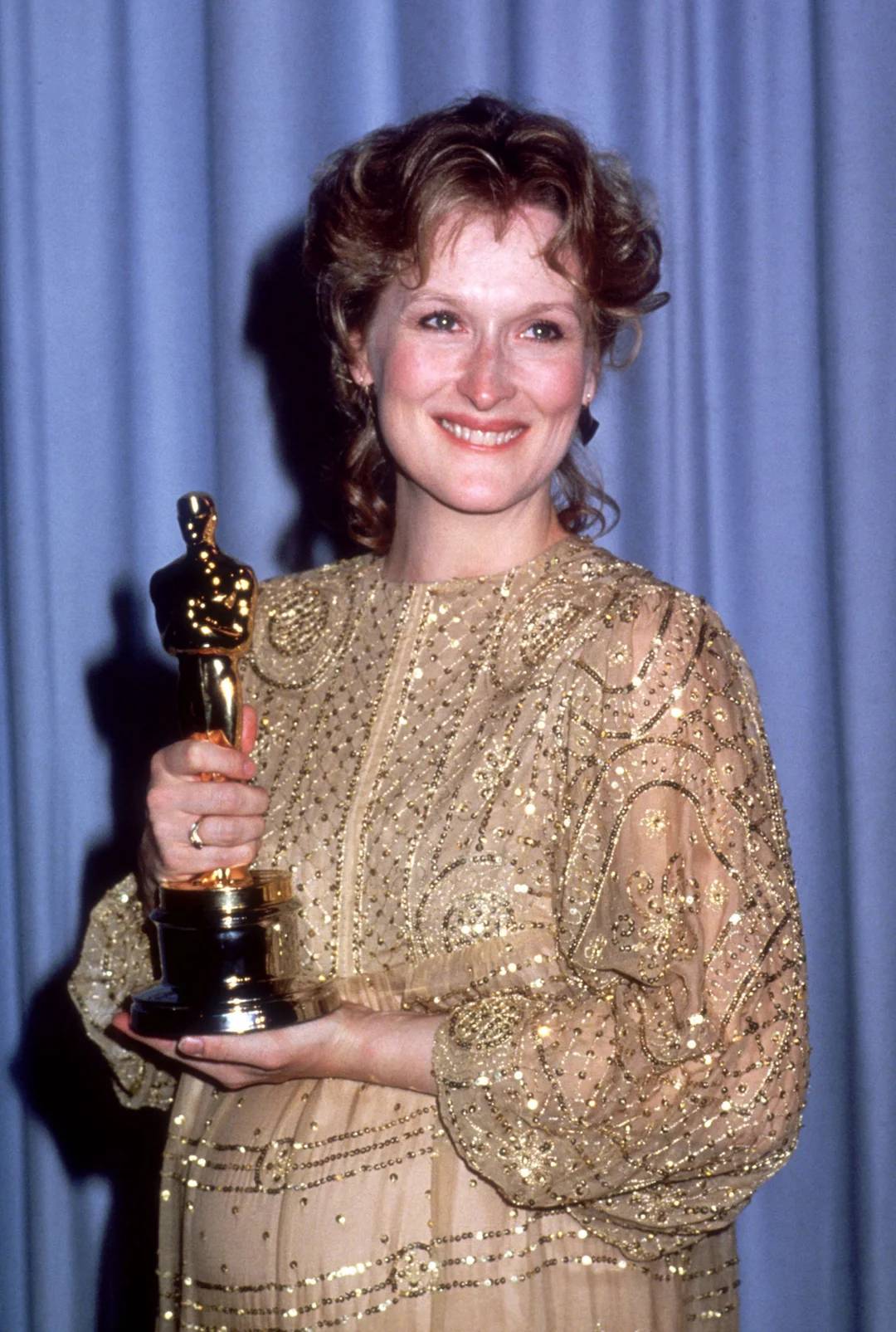A woman with wavy hair, wearing a sparkling, gold, long-sleeved dress, smiles while holding an Oscar statuette in front of a blue curtain.