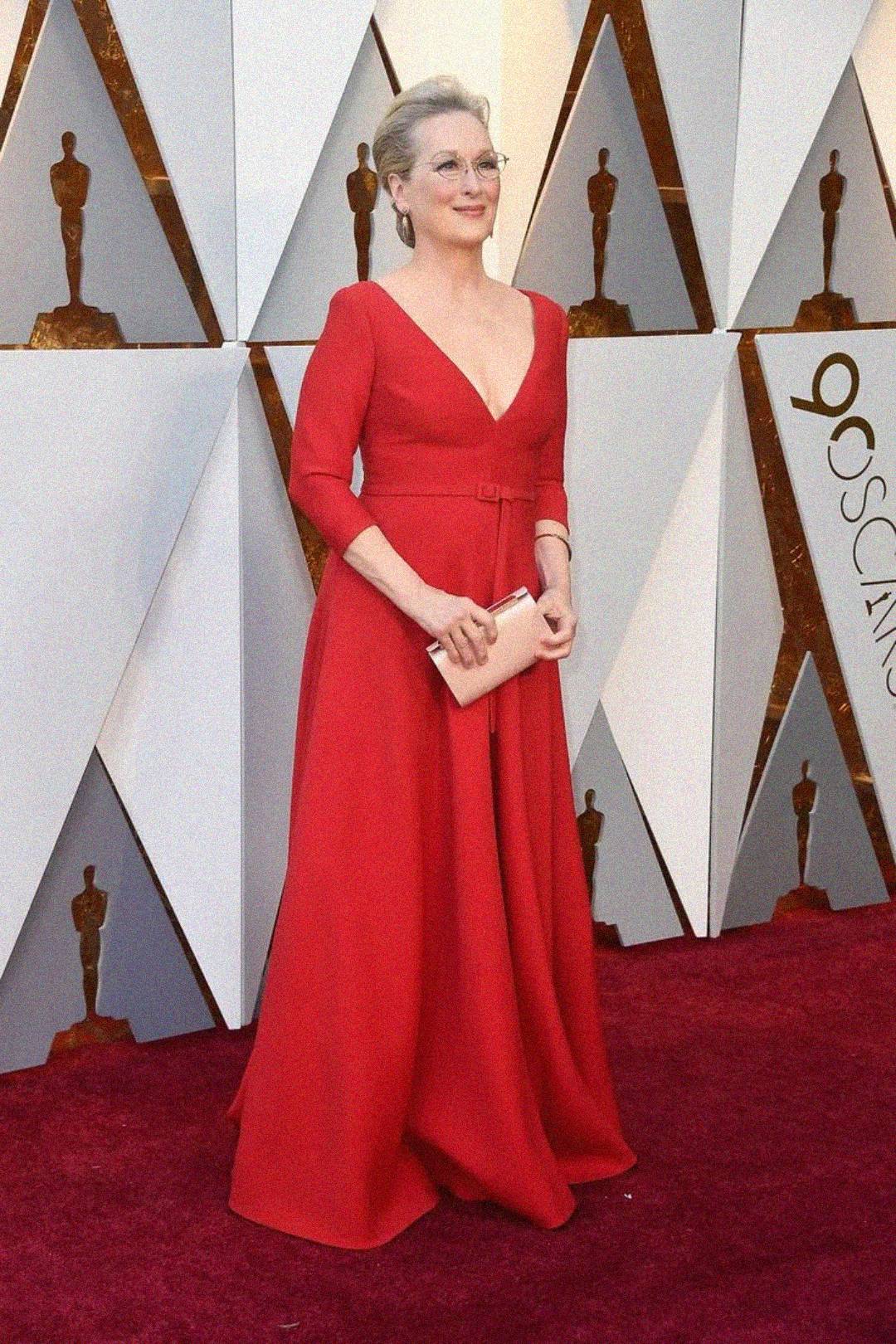 A woman in a red gown with a deep V neckline stands on the red carpet at the Oscars, holding a blush pink clutch. The background features Oscar statuette images and geometric panels with “90th Oscars” visible.