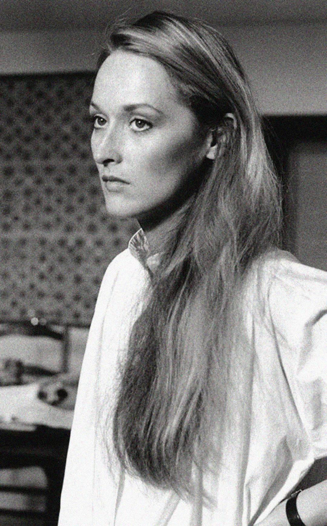 A black and white photo of a woman with long, straight hair wearing a light-colored shirt, looking thoughtfully to the side. The background is softly out of focus, showing a patterned wall and indistinct furniture.