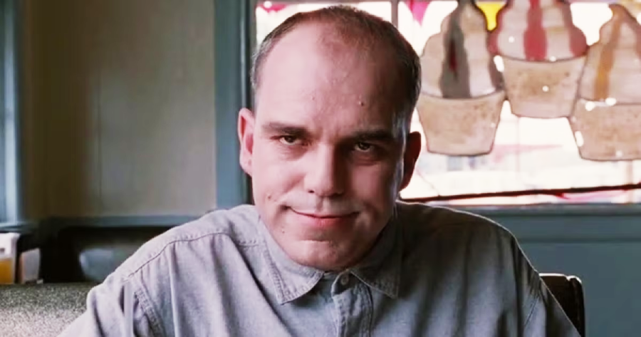 A man with a receding hairline and a slight smile sits in a diner, wearing a buttoned-up gray shirt. The window behind him has decorations shaped like ice cream cones.