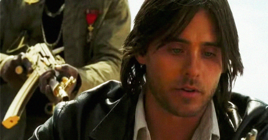 A man with long dark hair and a serious expression sits in the foreground, while another person behind him points a golden assault rifle in his direction. The background is bright and blurred.