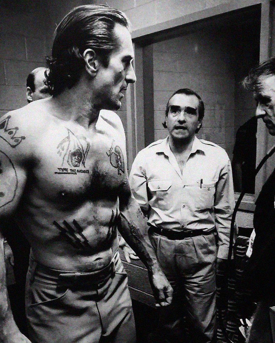 A shirtless man with tattoos stands in a room, facing two other men in conversation. One man wears a collared shirt and glasses; the setting appears to be backstage or on a film set, in black and white.
