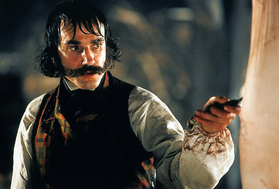 A man with dark hair and a thick mustache, wearing a plaid vest and white shirt with bloodstains, holds a knife and looks intently at something out of frame.