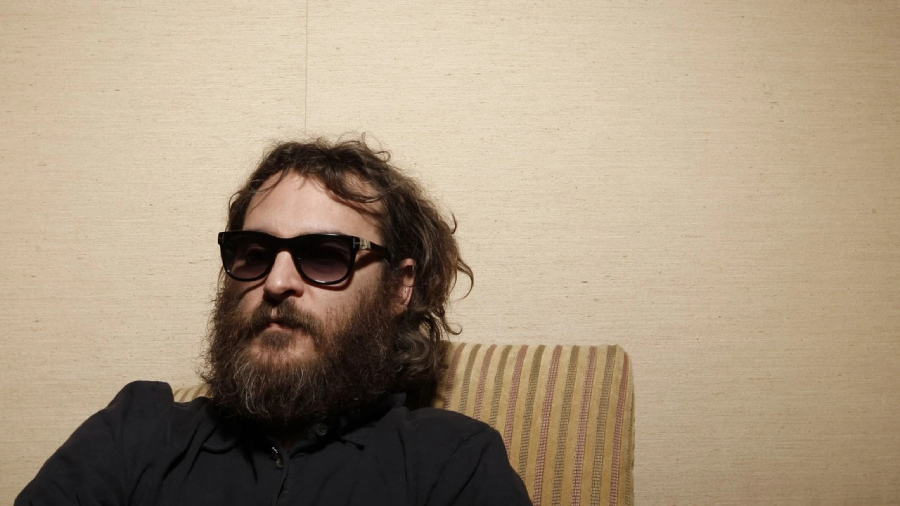 A man with a thick beard and long hair sits on a striped chair, wearing dark sunglasses and a black shirt, against a plain beige wall.