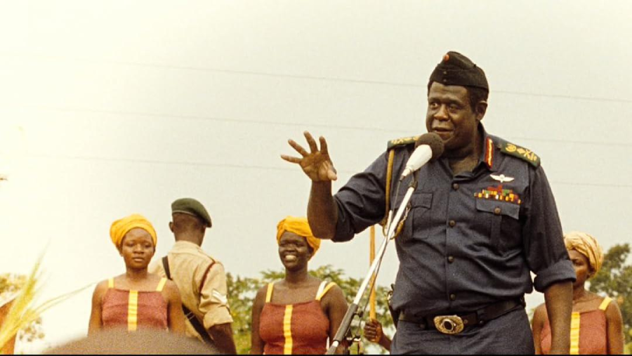 A man in a decorated military uniform speaks at a microphone outdoors, gesturing with his hand. Several women in matching yellow headwraps and dresses stand behind him, along with another uniformed person.