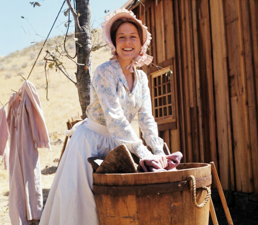 A woman dressed in pioneer-era clothing smiles while washing clothes in a wooden tub outdoors next to a wooden building. Dried grass and hanging laundry are visible in the background.