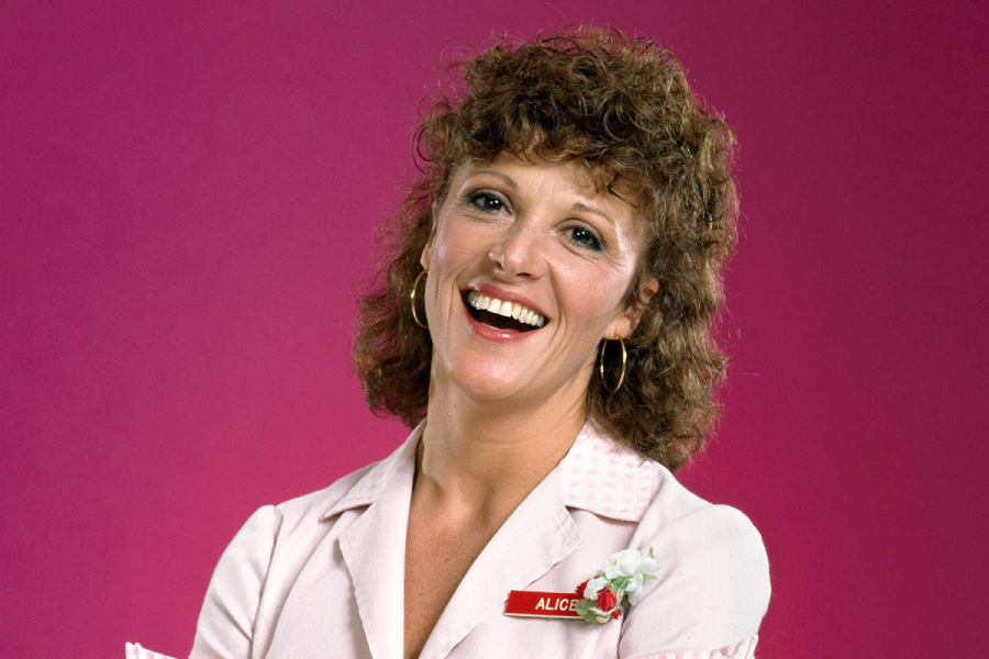 A smiling woman with curly brown hair, wearing gold hoop earrings and a light pink uniform with a name tag that reads "Alice," stands in front of a solid pink background.