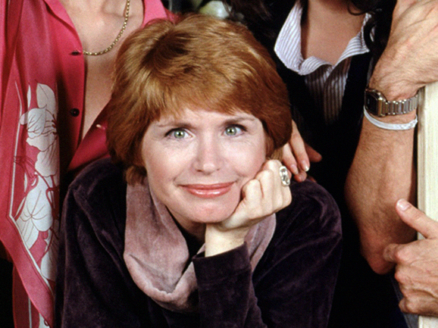 A woman with short red hair and a warm smile rests her chin on her hand. She wears a mauve scarf and dark top, with people partially visible around her.