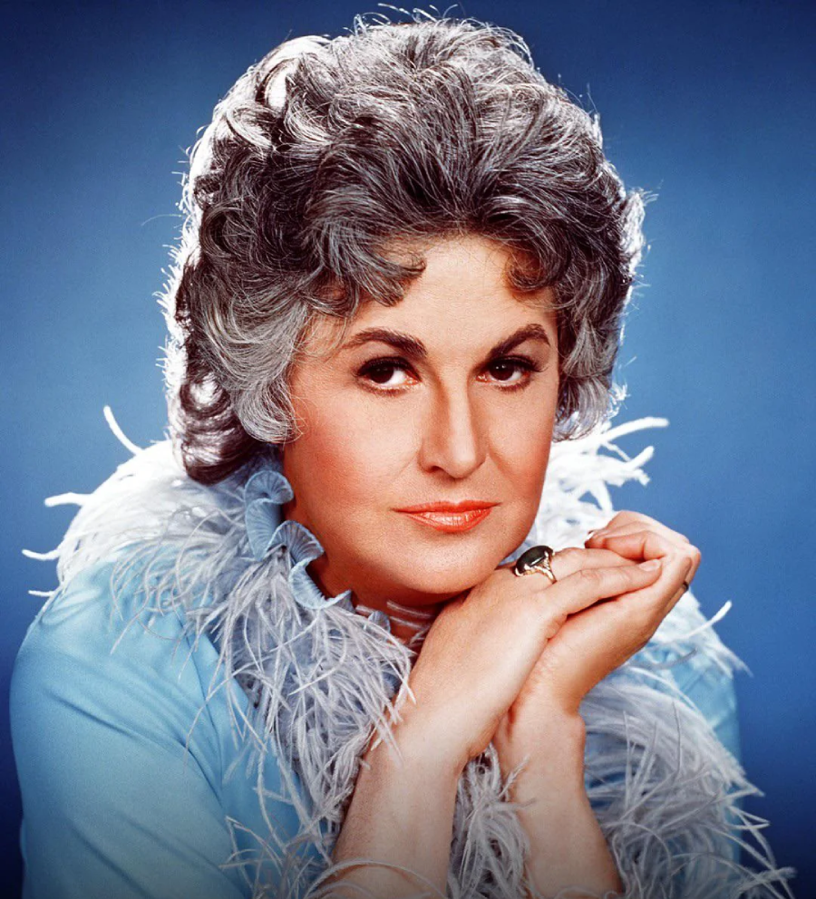 A person with short, wavy gray hair poses against a blue background, wearing a light blue feather-trimmed outfit and gently clasping their hands under their chin, looking confidently at the camera.