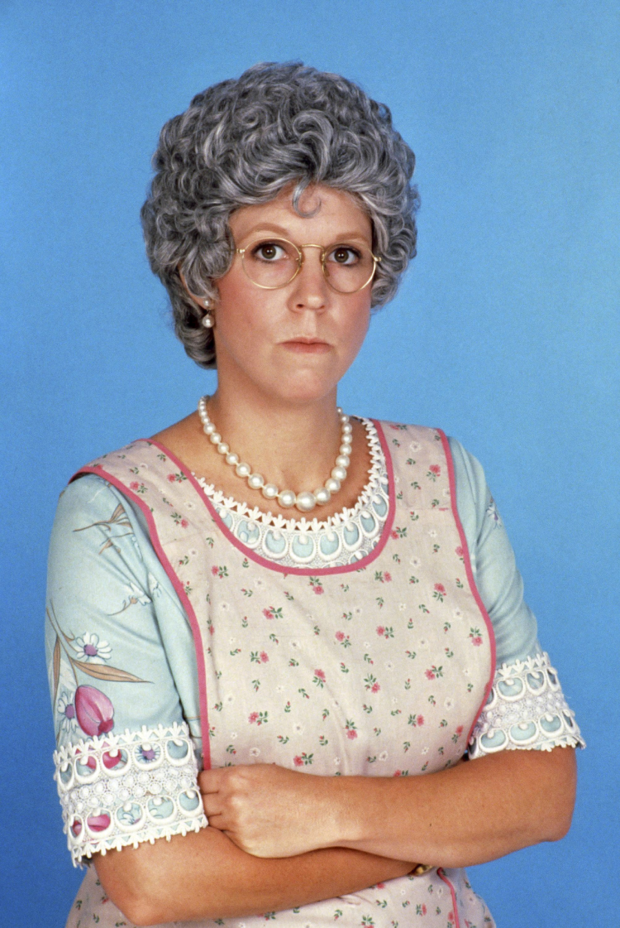A woman with curly gray hair, glasses, and a pearl necklace wears a floral dress and apron, standing with her arms crossed against a blue background. She has a serious expression.