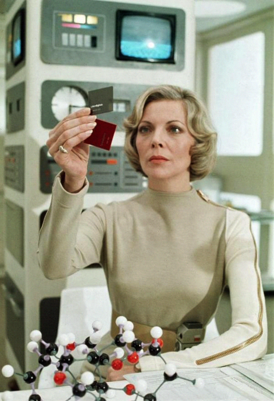 A woman in a futuristic outfit examines colored cards in a high-tech lab with control panels and molecular models on the table.