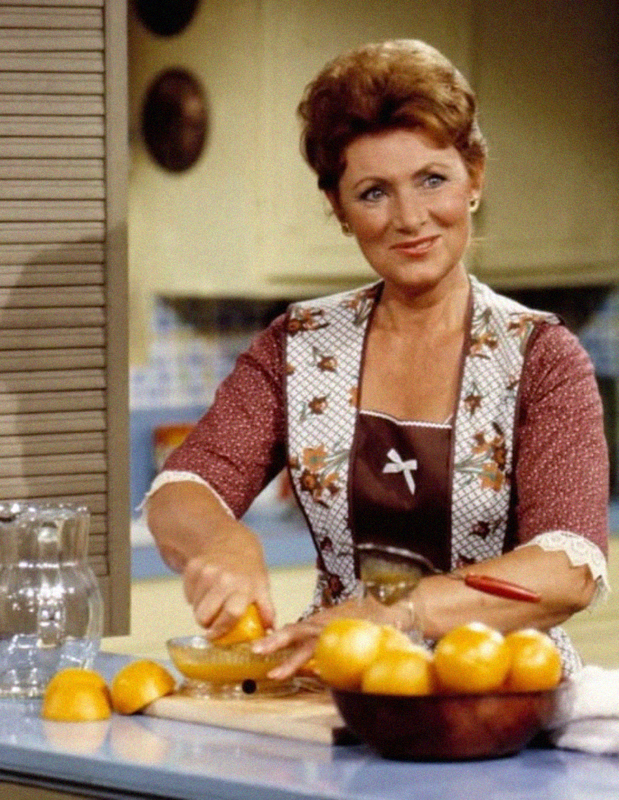 A woman in a patterned dress and apron stands at a kitchen counter, smiling as she squeezes oranges into a bowl. Several whole and halved oranges are on the counter beside a glass pitcher.