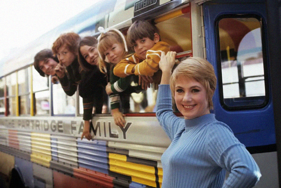 A smiling woman stands by a colorful bus as five children lean out of its windows, all smiling and looking at the camera. The bus is painted in bright, horizontal stripes.