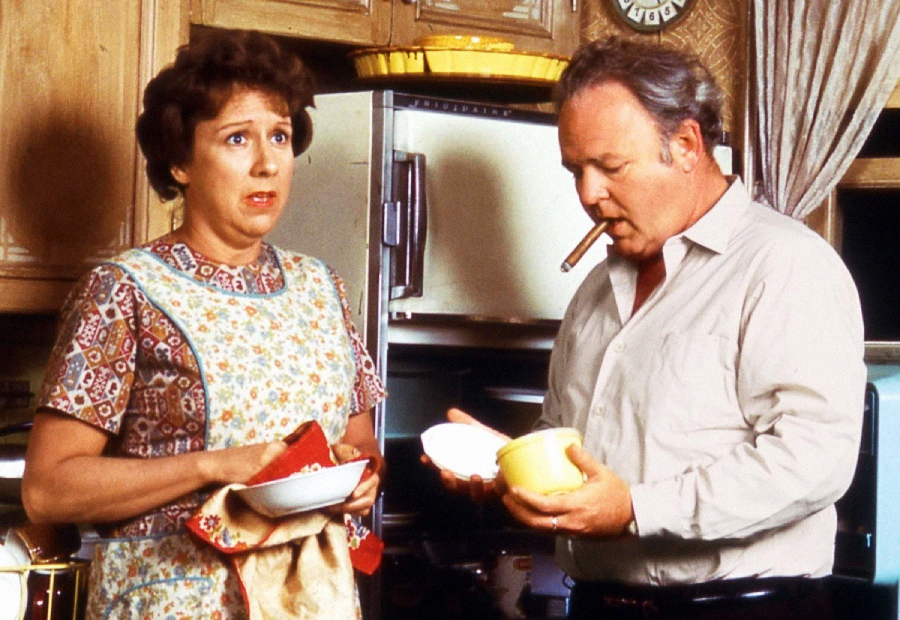 A woman in a patterned apron looks concerned while standing next to a man holding a cigar and dishes in a kitchen. Wooden cabinets and a refrigerator are visible in the background.