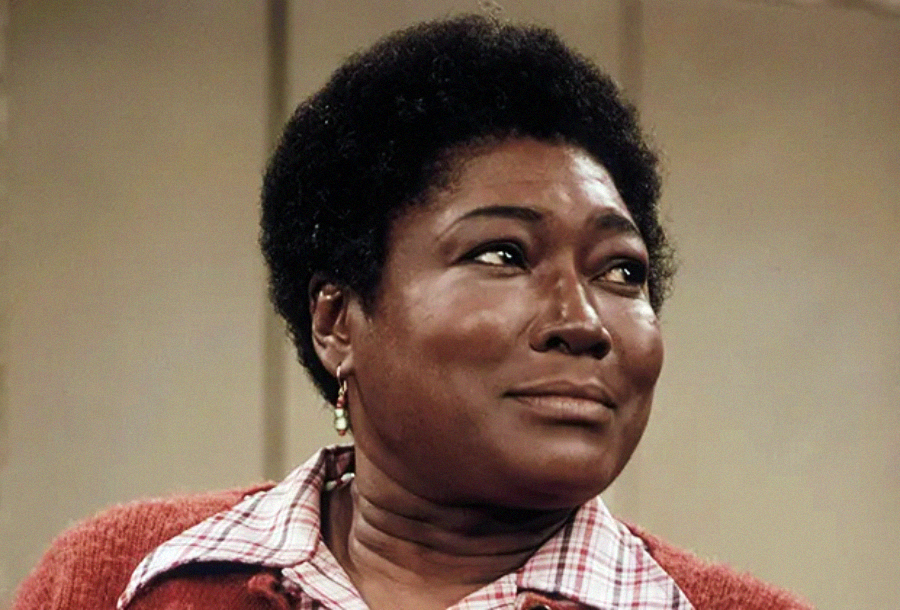 A woman with short curly hair and dark skin wears a plaid shirt under a red sweater. She looks off to the side with a thoughtful, emotional expression. The background is neutral and out of focus.