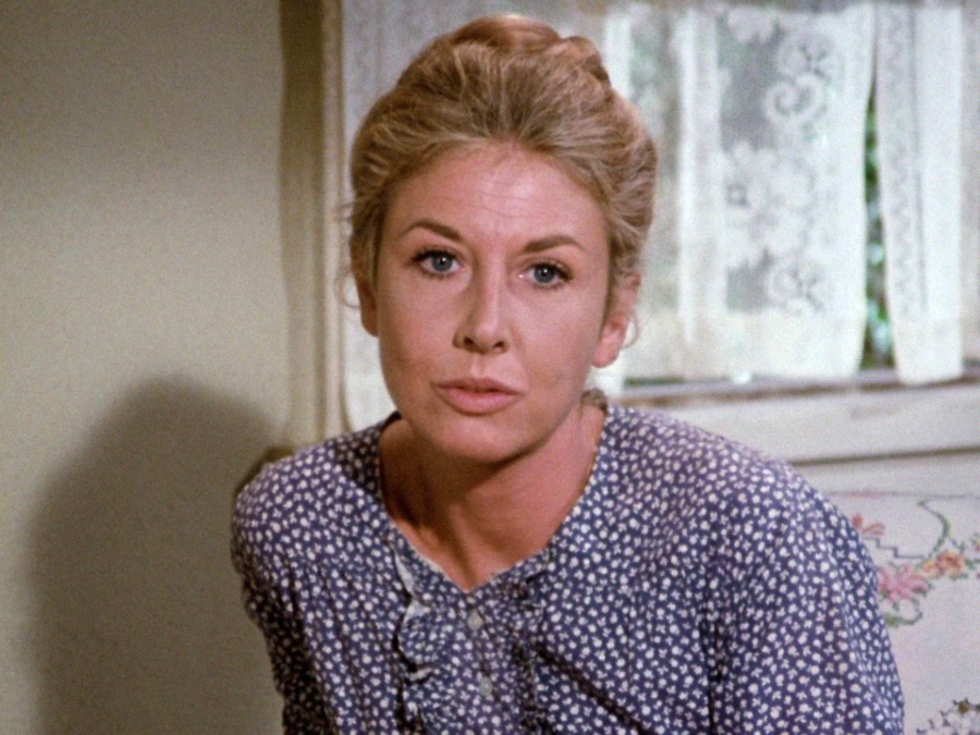 A woman with blonde hair tied back wears a blue patterned blouse and sits indoors near a window with lace curtains, looking slightly to the side with a neutral expression.