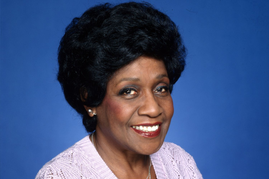 A smiling woman with short black hair, wearing a light purple sweater and pearl earrings, poses in front of a solid blue background.