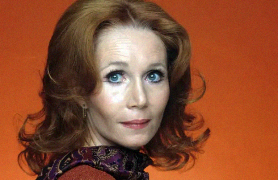 A woman with shoulder-length red hair and blue eyes looks over her shoulder against an orange background, wearing a patterned scarf and a rust-colored top.