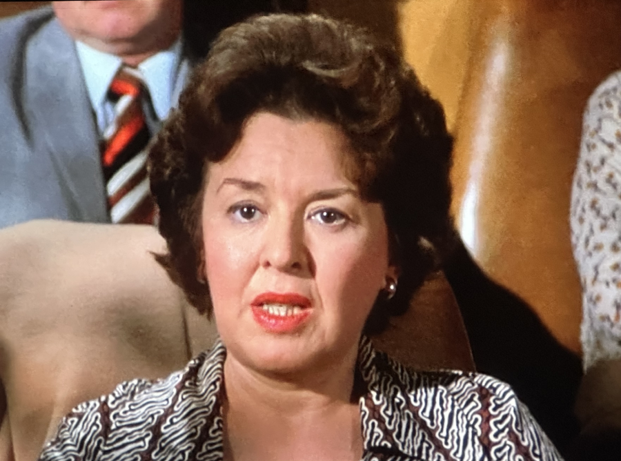 A woman with short curly brown hair and red lipstick speaks while seated, with two people partially visible behind her. She wears a patterned blouse and appears to be indoors, likely in a formal or public setting.