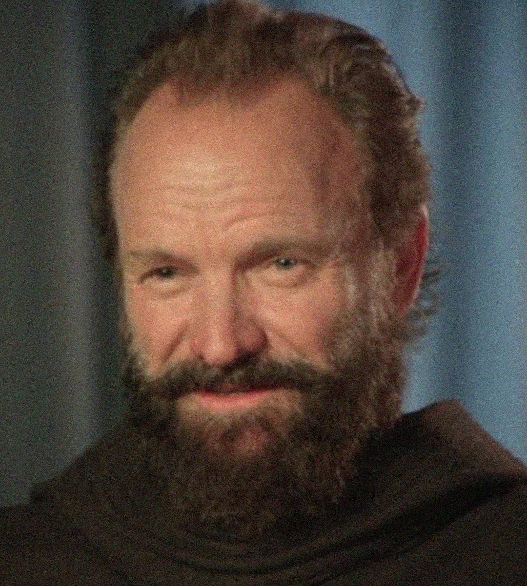 A man with a full brown beard and mustache smiles slightly. He has light skin, short hair, and is wearing a dark garment. The background is softly blurred in shades of blue and gray.