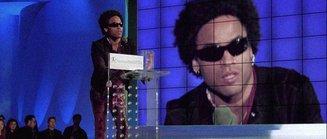 A person wearing sunglasses and a patterned outfit stands at a clear podium, speaking into a microphone, with their image displayed on a large screen behind them at an awards event.