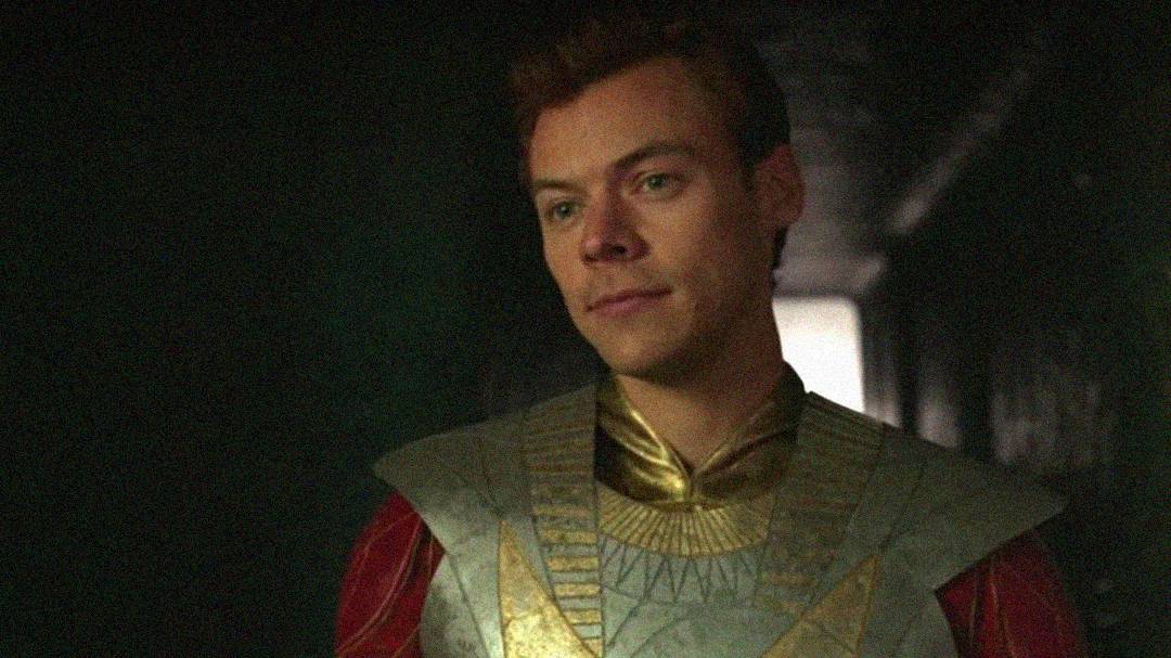 A person with short brown hair wearing a metallic, gold and silver futuristic costume with intricate patterns stands in a dimly lit hallway, looking ahead with a neutral expression.