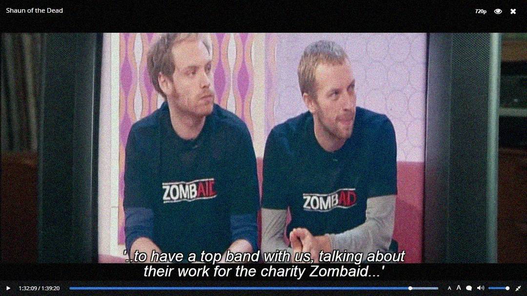 Two men wearing black "ZOMBAID" T-shirts sit side by side on a talk show set in the film "Shaun of the Dead." Subtitles read: "...to have a top band with us, talking about their work for the charity ZombAid...