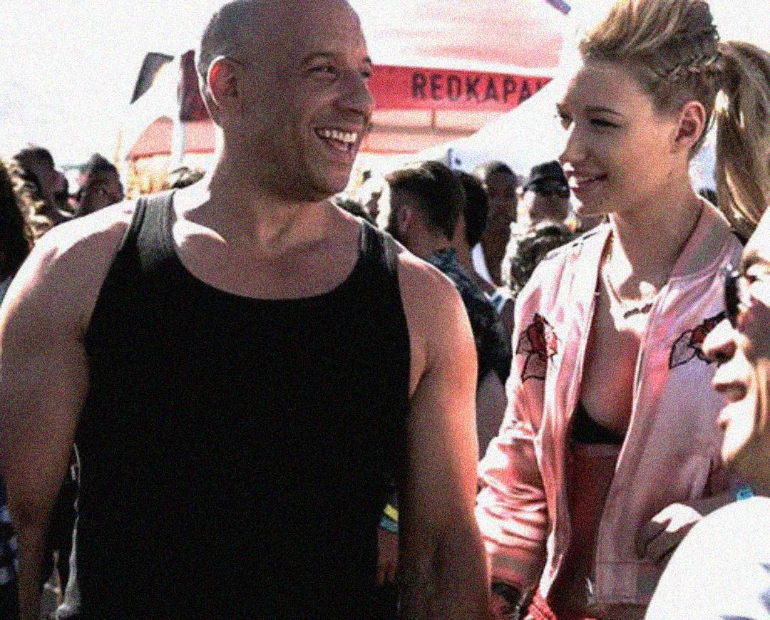 A muscular man in a black tank top smiles while standing next to a woman in a pink jacket at an outdoor event surrounded by a crowd. They appear to be enjoying themselves.