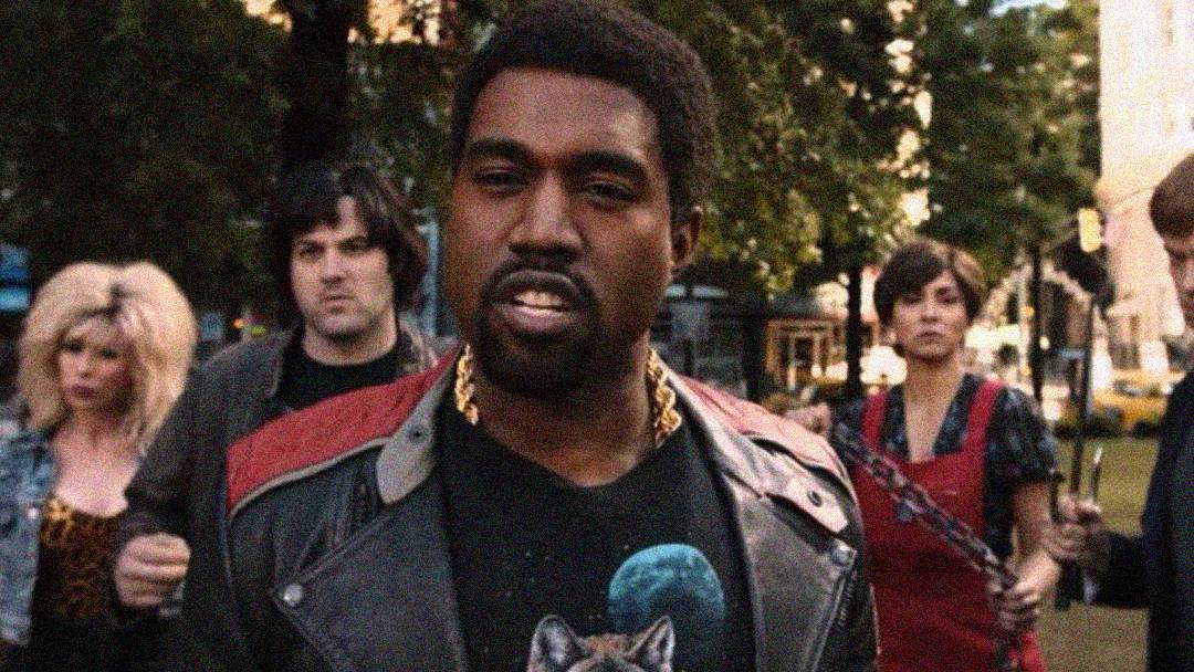 A man in a black leather jacket stands outside, looking forward, with three people behind him. Trees and buildings are visible in the background, and the group appears to be in a city park.