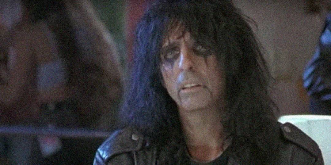 A person with long black hair and heavy black eye makeup wears a black leather jacket and looks forward with a serious expression. The background is blurred with indistinct figures and dim lighting.