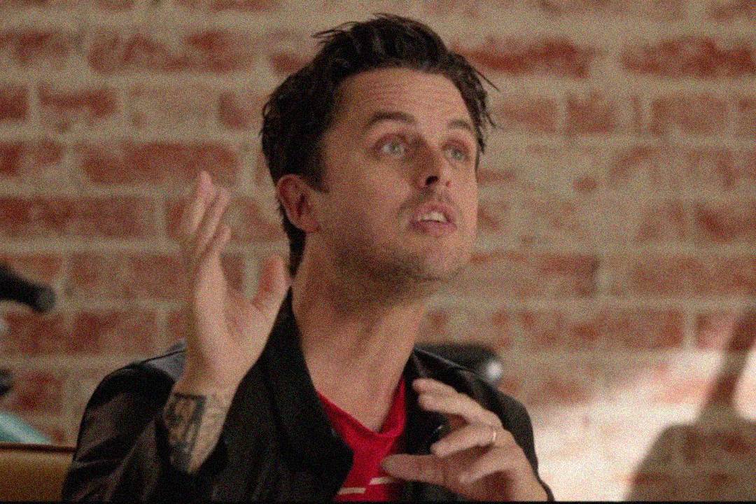 A person with short dark hair and a jacket gestures animatedly with their hands, looking upward. They are seated indoors in front of a red brick wall.
